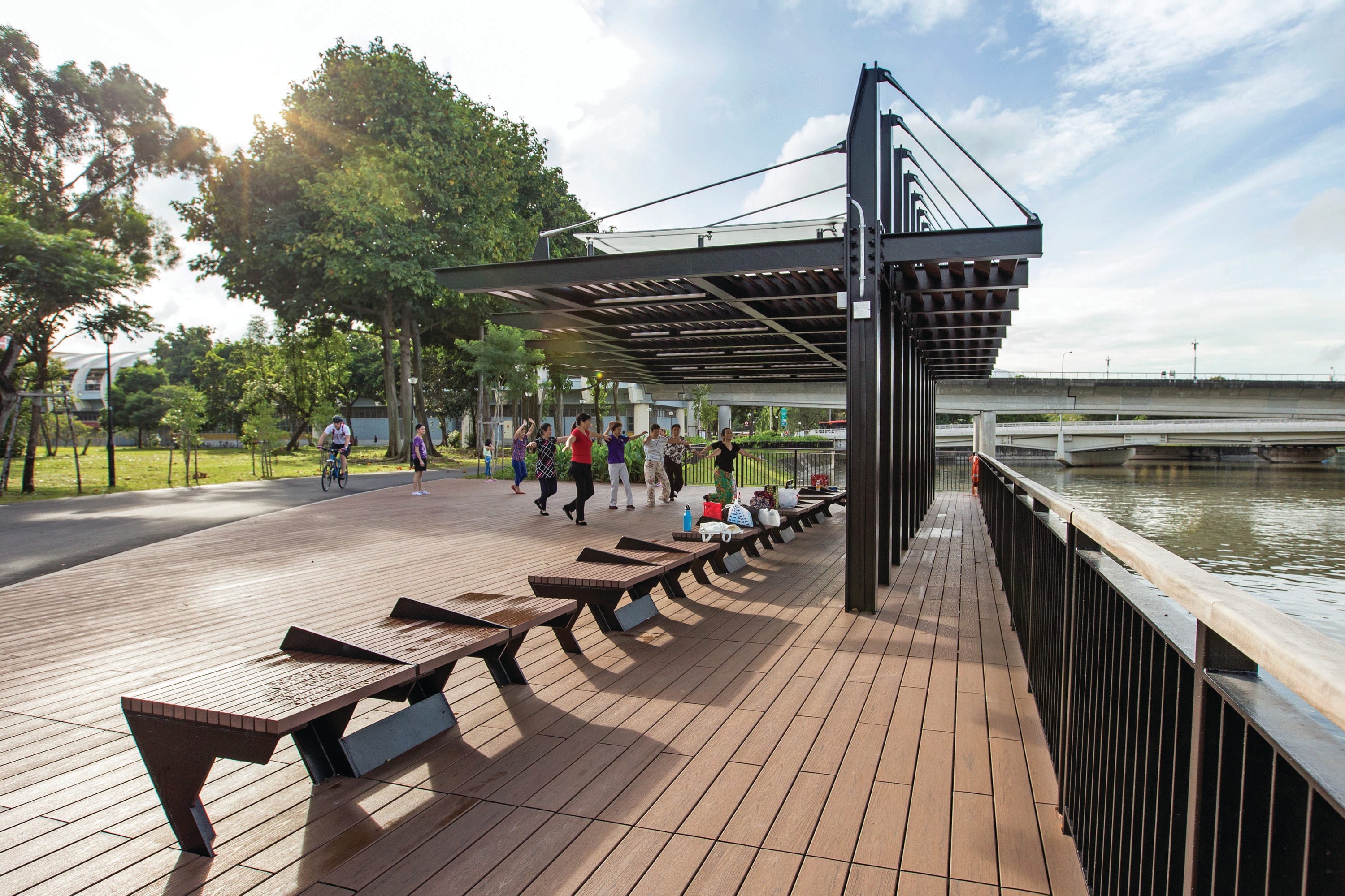 Public park with a wooden boardwalk, benches, and a metal pergola. People are exercising and cycling in the background. A river is visible on the right.